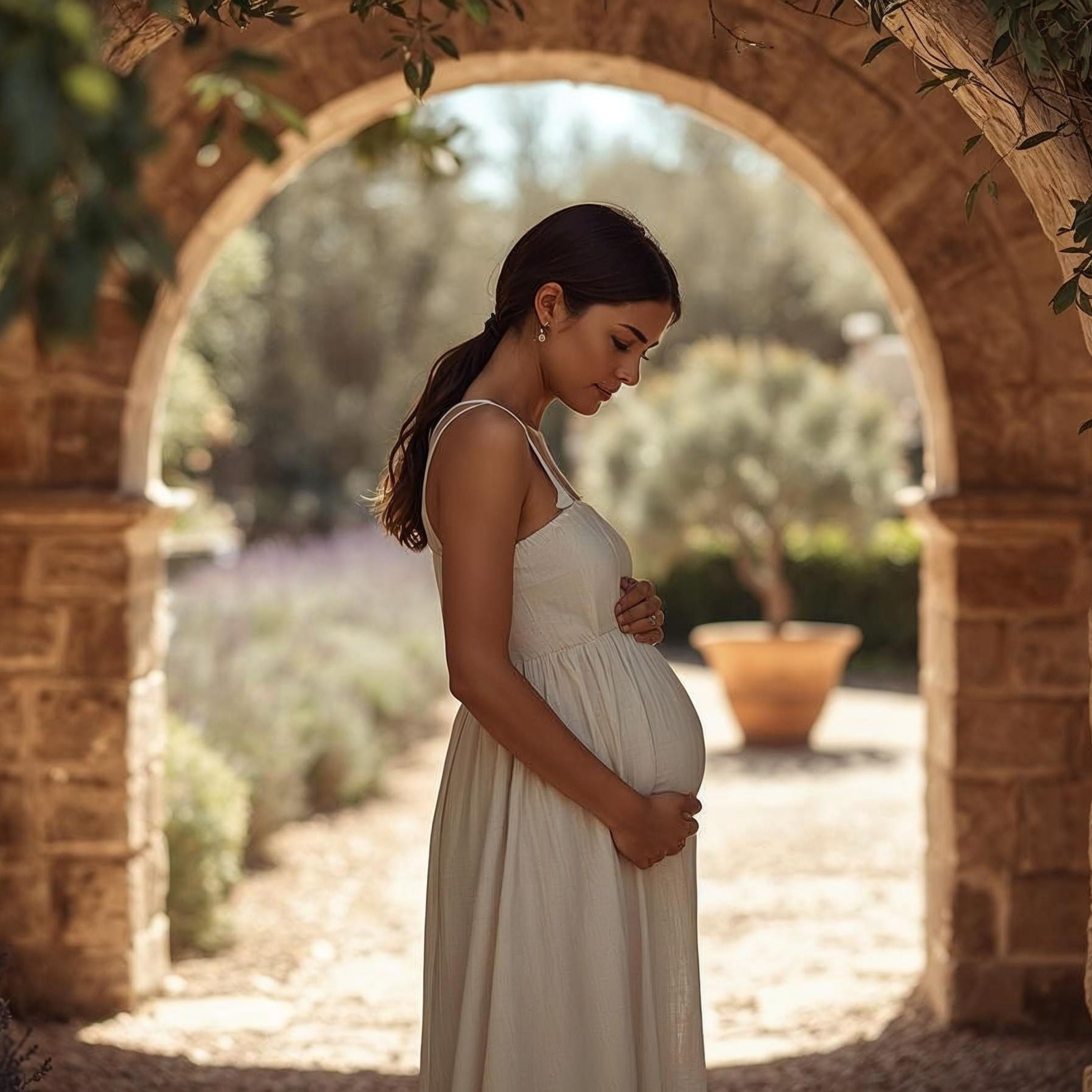 Pregnant woman in flowing cream dress cradles belly under stone arch, serene garden backdrop, natural light, fertility & pregnancy collection.